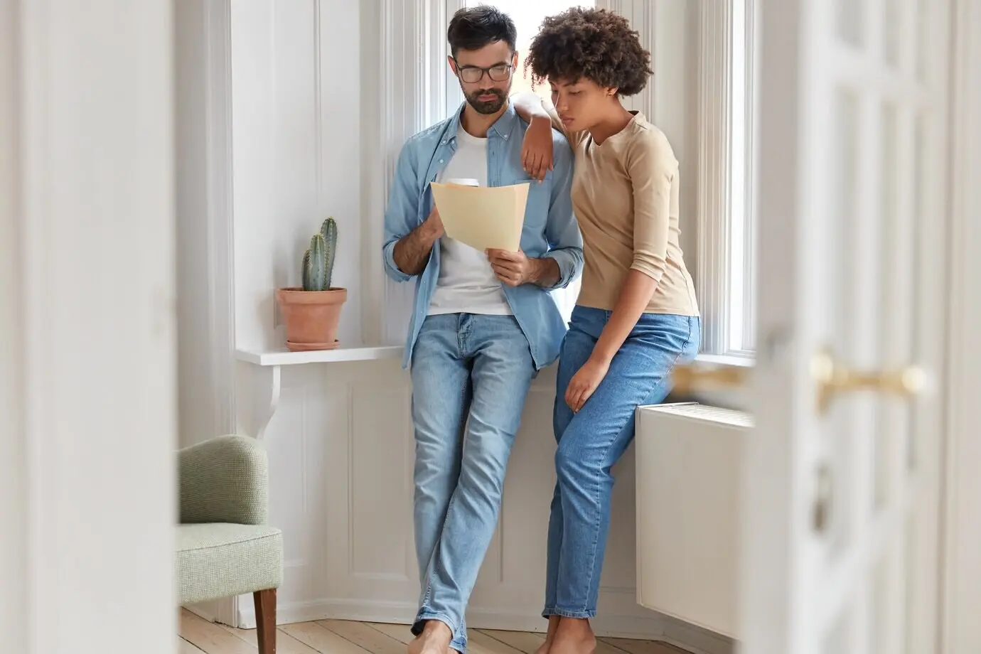 Een echtpaar bekijkt een rekening, plant hun budget en berekent hun uitgaven, is gekleed in jeans, drinkt koffie om mee te nemen en poseert in een modern appartement bij het raam. Twee interraciale partners bespreken zakelijke documenten.